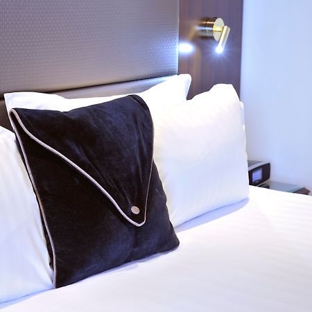 Stamford Plaza Melbourne Ξενοδοχείο Εξωτερικό φωτογραφία The photo shows a neatly arranged bed with white linens. There are several pillows, with one black pillow featuring a distinctive design on top of the white pillows. The headboard of the bed is visible, along with a stylish wall-mounted light fixture