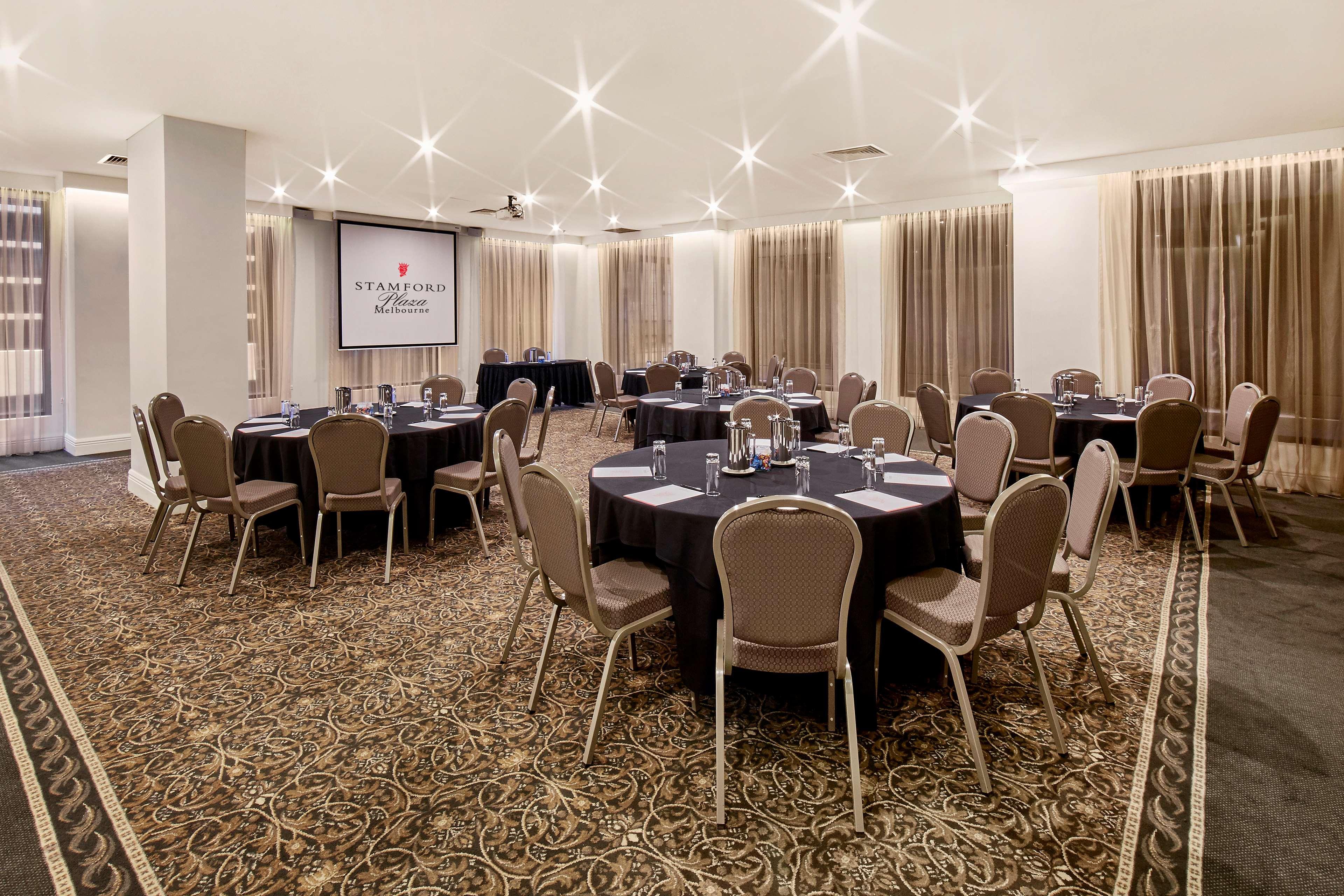Stamford Plaza Melbourne Ξενοδοχείο Εξωτερικό φωτογραφία The photo shows a well-lit event space set up for a gathering or meeting. There are several round tables covered with black tablecloths, each surrounded by beige chairs. In the background, a large screen displays a logo or branding, likely related to