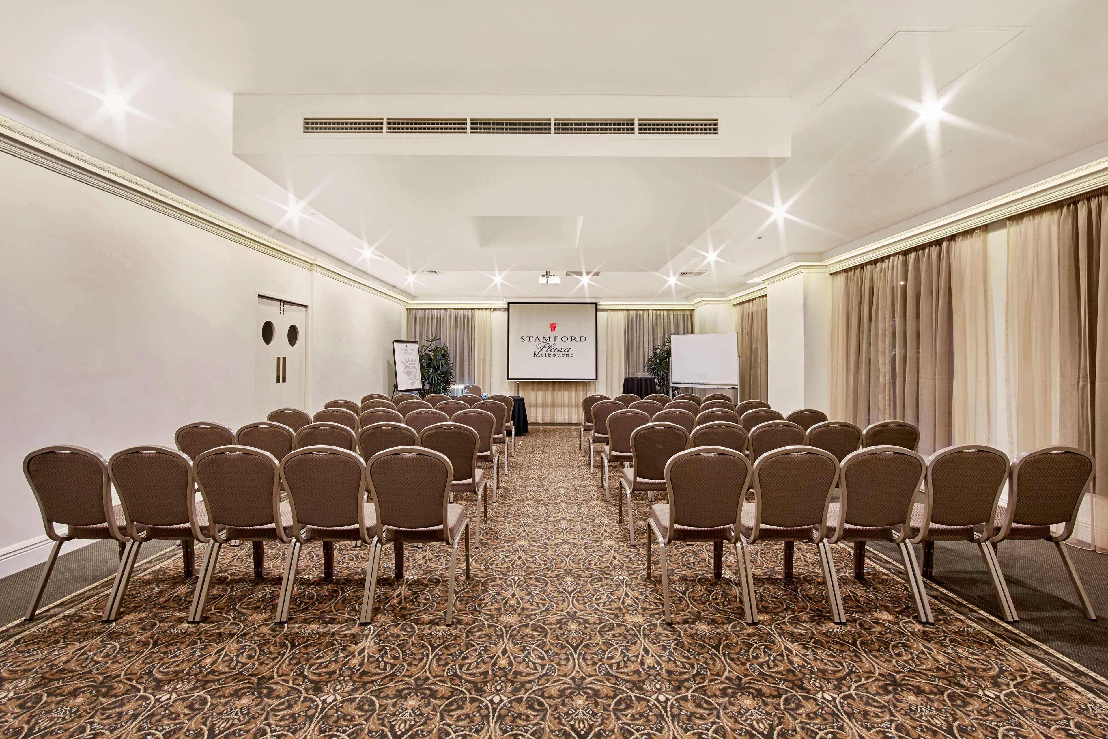 Stamford Plaza Melbourne Ξενοδοχείο Εξωτερικό φωτογραφία The photo shows a conference room set up for an event. It features several rows of beige chairs arranged facing a central presentation area. At the front, there is a screen displaying a logo or title, possibly for a conference or meeting. The walls h