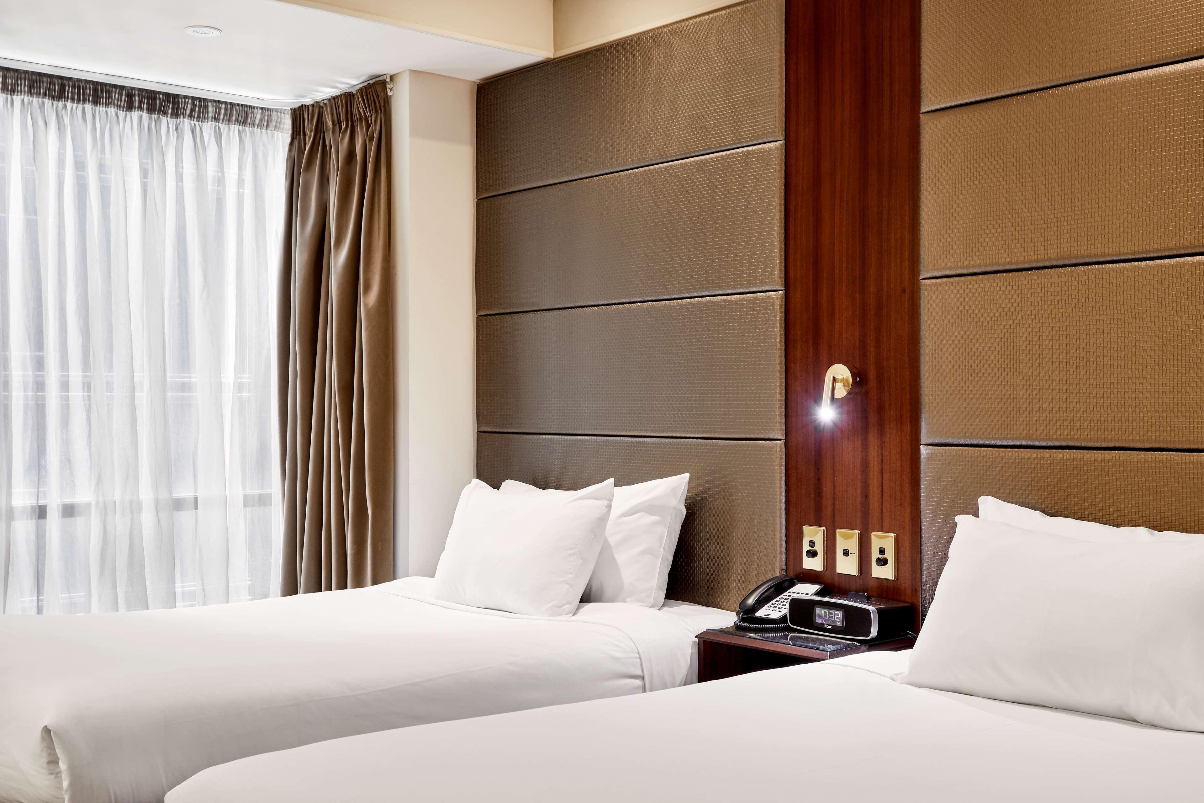 Stamford Plaza Melbourne Ξενοδοχείο Εξωτερικό φωτογραφία The photo shows a modern hotel room featuring two twin beds with white linens and fluffy pillows. The walls behind the beds have a combination of textured panels in neutral colors. There is a bedside table with a lamp and a phone. A large window with