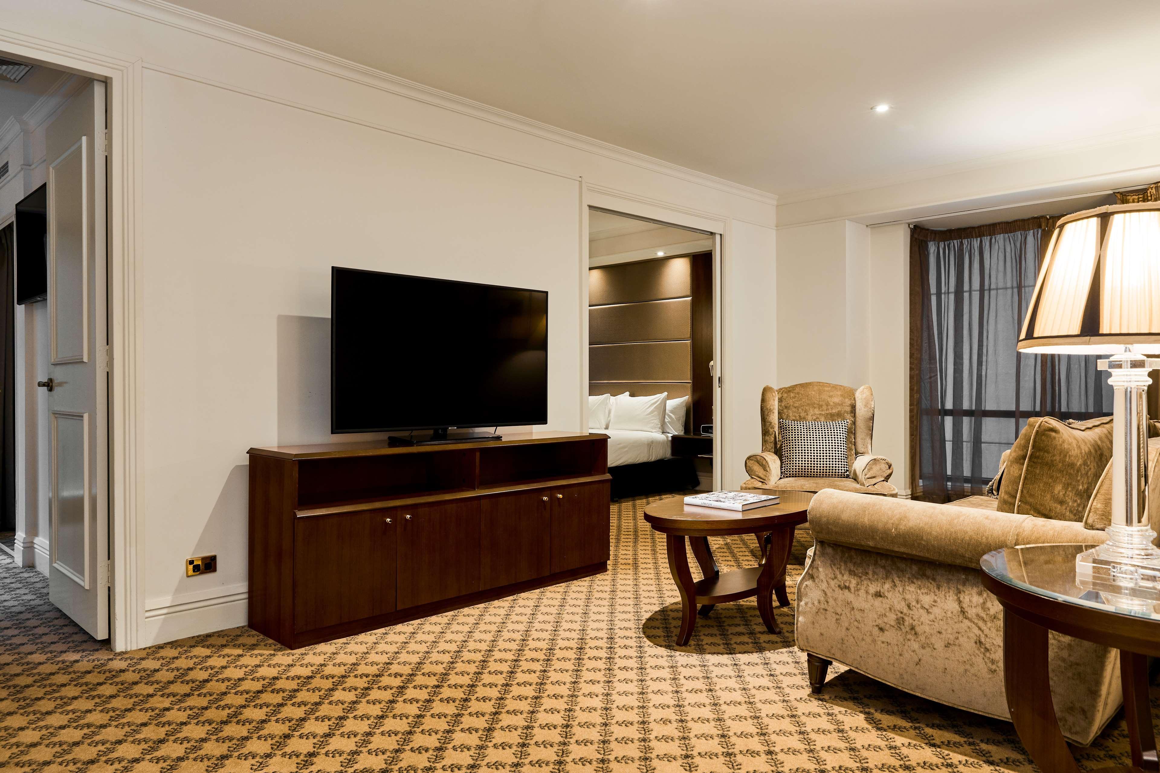 Stamford Plaza Melbourne Ξενοδοχείο Εξωτερικό φωτογραφία The photo shows a well-furnished room, likely a hotel suite or a living area. There is a large flat-screen television mounted on a wooden entertainment unit. In front of the TV, there is a round wooden coffee table. To the side, a comfortable armchai