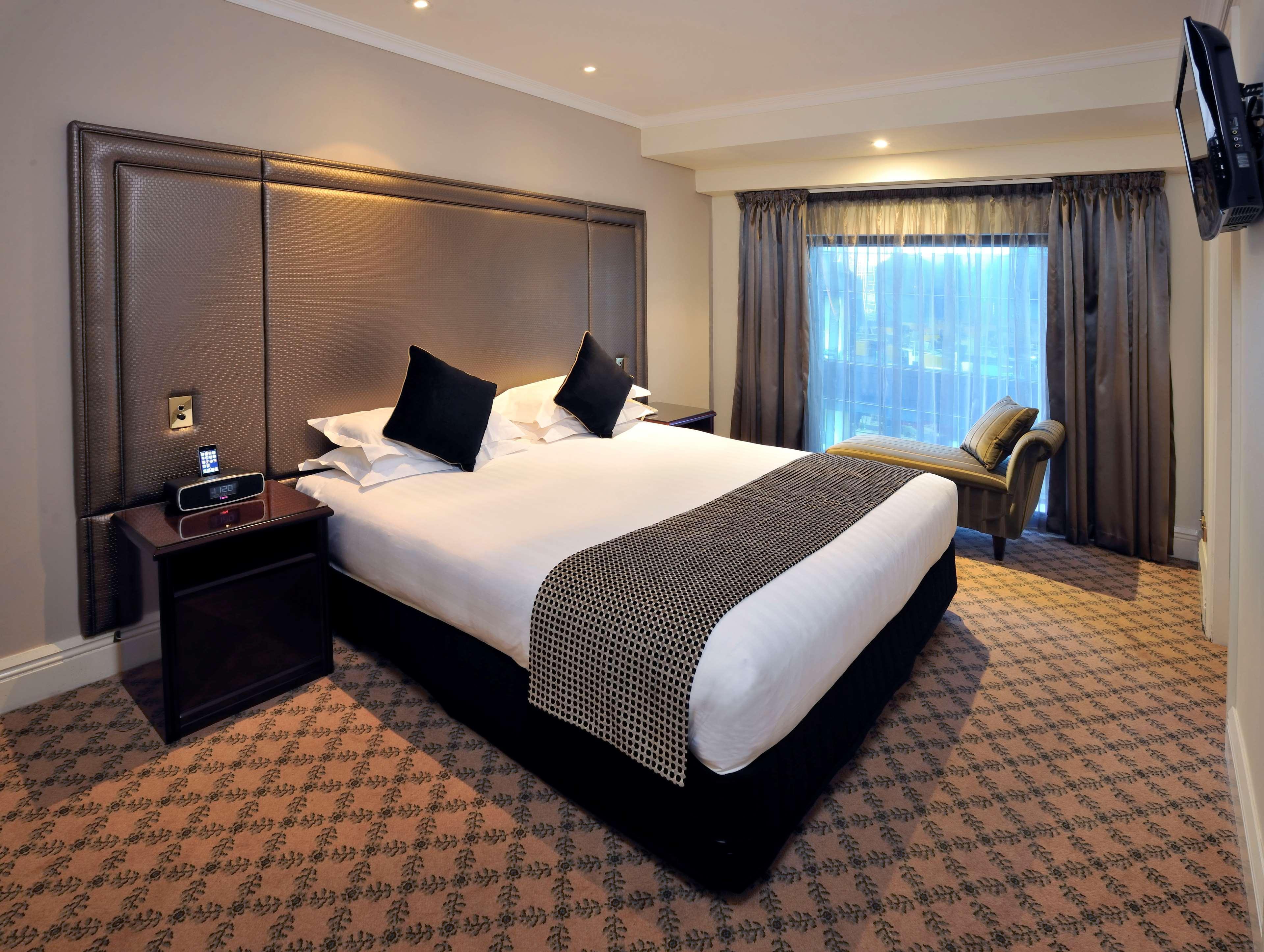Stamford Plaza Melbourne Ξενοδοχείο Εξωτερικό φωτογραφία The photo shows a hotel room featuring a neatly made king-sized bed with dark pillows. The headboard is upholstered in a dark material, and there is a decorative blanket at the foot of the bed. Beside the bed are two nightstands, each equipped with l